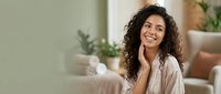 Young woman with curly dark hair and olive skin tone, wearing a light beige shirt, smiling while touching her face in a bright, plant-filled interior space.