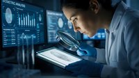 Young Asian male scientist examining data on tablet with magnifying glass in modern laboratory with digital analytics displays on background.
