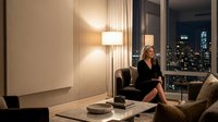 Professional blonde businesswoman in elegant black dress relaxing in luxury hotel suite with city skyline view at night for corporate travel content.