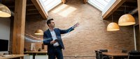 Professional businessman in dark suit presenting in modern loft office with exposed brick walls, wooden beams and pendant lighting for corporate presentations.