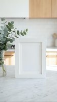White picture frame mockup on marble counter with eucalyptus branches in modern kitchen interior for photo display and home decor projects.