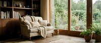 Cozy modern living room interior with comfortable beige armchair, built-in bookshelves, large windows overlooking lush garden, warm natural lighting for home decor.