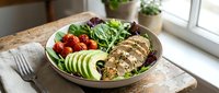 Nutritious grain bowl with grilled chicken breast, fresh vegetables, and leafy greens arranged on wooden table near window for healthy eating and meal prep.