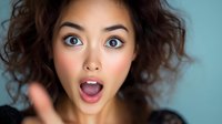 Young Asian woman with curly hair expressing surprise and excitement with open mouth and wide eyes against blue background. Emotional portrait showing genuine amazement.