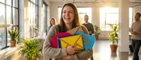 Young woman with long brown hair and light skin tone, wearing casual beige cardigan, holding colorful envelope clutch in office environment with colleagues and plants.