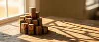 Natural wooden building blocks stacked on floor with warm sunlight streaming through window creating geometric shadows on hardwood surface.