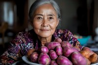 Senior Asian woman with gray hair smiling warmly while holding fresh purple potatoes on plate, wearing floral pattern dress, representing healthy aging and traditional farming lifestyle.