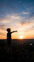 Child silhouette pointing at golden sunset sky from hilltop, exploring nature and landscape during evening hour for travel and adventure concepts.