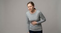 Young Caucasian woman experiencing stomach pain holding abdomen in grey sweater against neutral background for health and medical concepts.