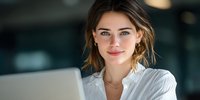 Young professional woman with brown hair smiling confidently while working on laptop in modern office environment, showcasing workplace positivity.
