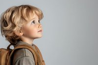 Young child with curly blonde hair wearing backpack looking up with wonder and curiosity against gray background, profile view portrait capturing innocent expression.