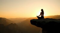 Woman meditating in lotus pose on mountain cliff edge during golden sunset with layered mountain ranges creating peaceful spiritual atmosphere.