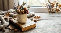 Cozy autumn still life with dried flowers in ceramic mug on wooden table near window. Warm seasonal home decor with knitted blanket and natural elements.