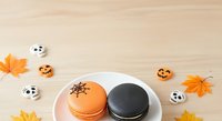 Colorful Halloween macarons with ghost and pumpkin designs on white plate, surrounded by autumn leaves and seasonal decorations on wooden surface.