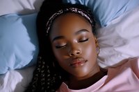 Young African American woman sleeping peacefully on white bedding, wearing pink pajamas and patterned headband, with long braided hair and natural makeup.