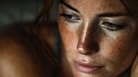 Close-up portrait of young woman with freckles and red hair in soft, dramatic lighting, showcasing natural beauty and thoughtful expression.