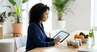 Professional African American businesswoman using tablet computer at modern kitchen table with healthy breakfast and green plants in bright home office.