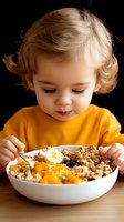 Adorable toddler with blonde curly hair eating healthy breakfast bowl with granola, fruits and nuts in yellow shirt against dark background.