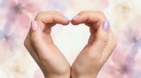 Two hands forming a heart shape against a soft, blurred floral background with pink and white flowers, expressing love, care, and affection.