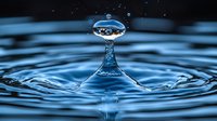 Macro photography of water drop collision creating crown splash and floating droplet above rippled blue water surface. Frozen motion captures liquid dynamics.
