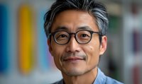 Mature Asian man with gray hair and glasses wearing denim shirt smiling warmly at camera against blurred colorful background. Professional headshot style portrait.