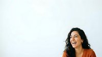 Young woman with long dark wavy hair and medium skin tone wearing an orange shirt, smiling and looking upward against a white background for portrait and lifestyle use.