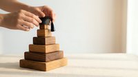 Hand placing wooden figure on top of pyramid made of stacked wooden blocks representing hierarchy, leadership development and team building concepts.
