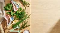 Fresh herbs and colorful measuring spoons arranged on wooden cutting board for cooking and baking preparation with natural kitchen styling and copy space.