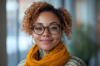 Young African American woman with curly hair and glasses wearing orange scarf smiling confidently in modern office environment for professional headshot.