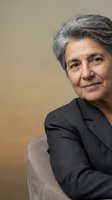 Senior woman with gray hair wearing dark blazer, seated and smiling warmly at camera against neutral beige background. Corporate portrait for business, leadership, healthcare, and professional services.