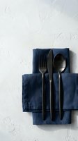 Overhead view of black cutlery set with fork, knife and spoon arranged on folded navy blue linen napkins against white textured background, suitable for restaurant, dining and tableware design projects.