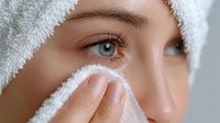 Close-up of Caucasian woman with blue eyes applying skincare treatment with white towel wrapped around head during spa facial routine.