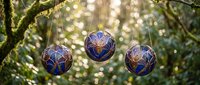 Three decorative blue and gold ornamental baubles hanging from tree branches with warm bokeh lights creating magical outdoor holiday atmosphere.
