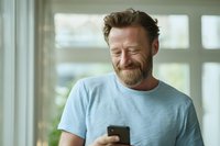 Middle-aged Caucasian man with beard smiling at smartphone in bright room with natural light, expressing joy while reading message or social media.