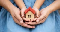 Family hands protecting wooden house model symbolizing home insurance, real estate investment and property security concept on blue background.