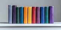 Row of colorful hardcover books standing in line on white shelf against neutral background, spines showing vibrant spectrum of colors from purple to orange.