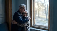 Senior Caucasian couple embracing by window in cozy home interior, enjoying peaceful moment together during golden hour with warm natural lighting.