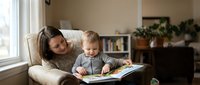 Young mother and toddler son reading book together on cozy chair by window in warm living room. Family bonding time, early childhood education concept.