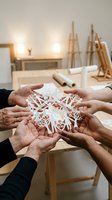 Multiple hands holding intricate white paper snowflake cutout in bright workshop studio with wooden easels and design tools, showcasing collaborative craftsmanship and seasonal decoration creation process.