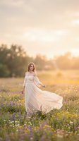 Young woman with long blonde hair wearing flowing white off-shoulder gown standing in blooming wildflower meadow at golden hour sunset. Romantic maternity or bridal portrait for wedding, maternity, fashion, and lifestyle photography.