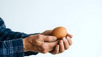 Close-up of mature male hands holding single brown egg against white background, suitable for cooking, nutrition, and food-related design projects.