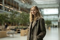 Young blonde businesswoman in checkered coat standing confidently in modern office lobby with natural lighting and contemporary architecture.