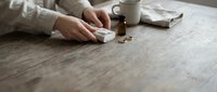 Woman taking medication pills from bottle on wooden table with coffee cup in cozy home setting for health and wellness routine.