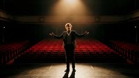 Young male performer standing on theater stage with arms outstretched facing empty red velvet seats under dramatic spotlight for acting audition rehearsal.
