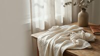 Cozy bedroom interior with unmade bed, soft natural lighting through sheer curtains, neutral beige linens, wooden furniture and dried eucalyptus branches in vase.