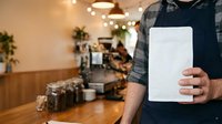 Barista in checkered shirt holding blank tablet displaying menu or promotional content in modern coffee shop with warm lighting and wooden counter.