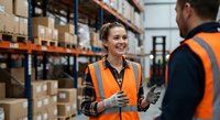 Smiling female warehouse worker in safety vest discussing logistics with male colleague in modern distribution center with shelving and boxes.