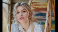 Young Caucasian woman with blonde wavy hair and natural makeup wearing white blouse poses outdoors during golden hour, soft dreamy portrait with warm lighting and bokeh effect.