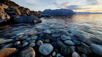 Dramatic coastal landscape with smooth stones and boulders along rocky shore at sunset, mountain silhouettes on horizon, calm ocean water reflecting golden light.