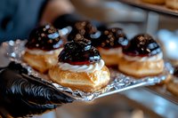Fresh cream puffs filled with whipped cream and topped with dark cherries and chocolate glaze on silver serving tray, served by hands in black gloves.